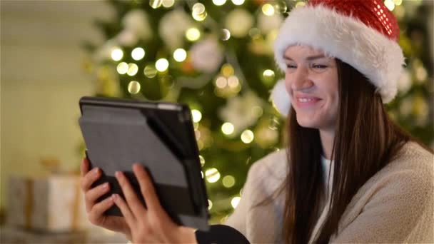 Gorgeous Lady in Santa Hat Calls Somebody Using the Video Camera on the Tablet During Celebration Christmas on Xmas Tree Background. - Felvétel, videó