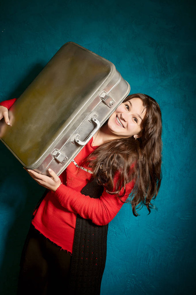 young brunette woman holding a big old suitcase against the backdrop of a blue wall - Photo, Image