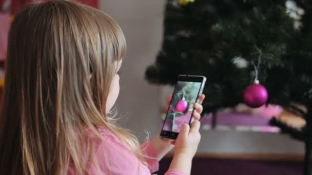 meisje foto's op een mobiele telefoon mooie kerstboom met feestelijke verlichting in de woonkamer in familiehuis - Video