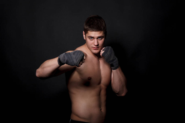 Hombre atlético muscular boxeando sobre un fondo negro. Un hombre con un torso desnudo en pantalones cortos, manos peremotanye vendajes para el boxeo
. - Foto, Imagen