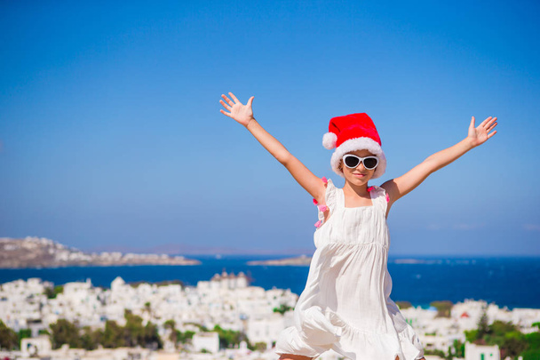 Kis vicces lány piros Santa kalap szabadban háttér-Mykonos. Gyerek Street, tipikus görög hagyományos falu, fehér falakkal és színes ajtók a karácsonyi Nyaralás Görögországban - Fotó, kép