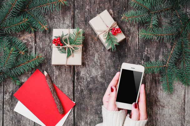 Női kezei mobiltelefon fa háttér a karácsonyi ajándékokat, fenyő ágak. Karácsonyt és boldog új évet összetétele. Lapos feküdt, top view - Fotó, kép