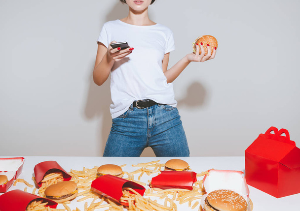 Closeup of young woman with burger standing and using smartphone - 写真・画像