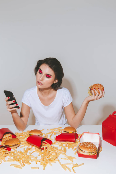 Pretty young woman with hamburger sitting and using mobile phone - Foto, afbeelding