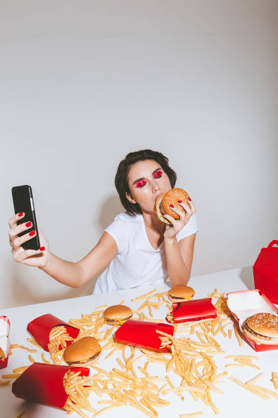 Attractive woman eating burger and taking selfie with cell phone - Foto, Imagen