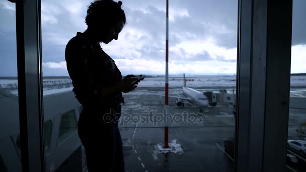 Sziluettjét fit kislány airport ablak közelében, és használ a smartphone. Formás, fiatal üzleti nő várja őt bentlakásos közelében a kapu az ablak és a gépet a háttérben. - Felvétel, videó