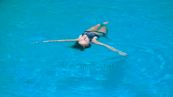 Adorable fit woman in black swimsuit is relaxing luying on the water. Young shapely girl is showing comfort and pleasure floating in the swimming pool. - Video, Çekim