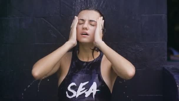 Cute girl standing under the shower outdoors. Girl in swimsuit stands in dark gray wall. It is heated under warm water, then to continue diving in cold water. - Footage, Video