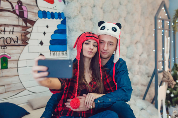 Beautiful young couple in funny hats sitting on the background of Christmas decorations and make selfie. - Fotoğraf, Görsel
