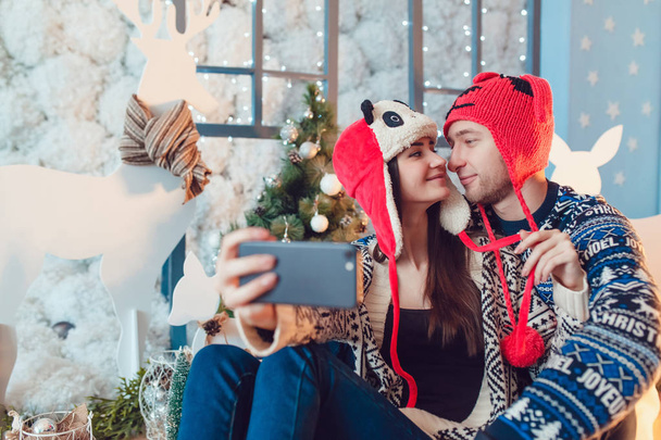 Beautiful young couple in funny hats sitting on the background of Christmas decorations and make selfie. - Valokuva, kuva
