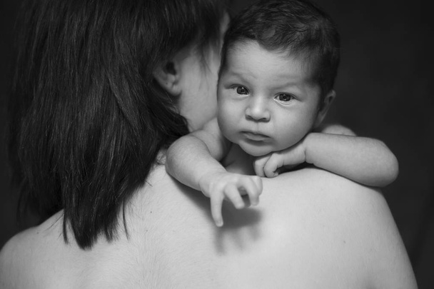 Little infant baby boy sleeping laying on mothers arms. Focus on a boy. Neutral black background, black and white picture. All in white clothes. Happy family - Foto, Bild