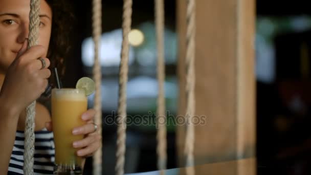 attraktive lächelnde Brünette spielt abends auf Schaukeln mit Cocktail in ihren Händen. junges schönes Mädchen trinkt Erfrischung und genießt ihren exotischen Urlaub in Indonesien. - Filmmaterial, Video