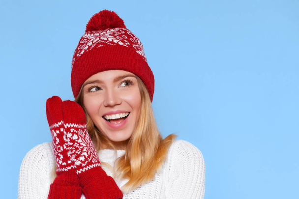 Surprised happy beautiful woman looking sideways in excitement. Christmas girl wearing knitted warm hat and mittens, isolated on blue background - 写真・画像