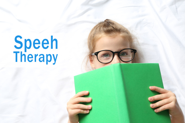 Little girl in glasses with book on bed. Text SPEECH THERAPY on background - Photo, Image