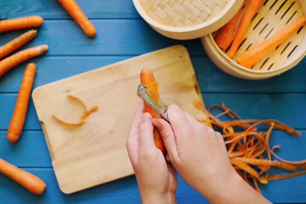 Женские руки чистят оранжевую морковку на синем фоне
 - Фото, изображение
