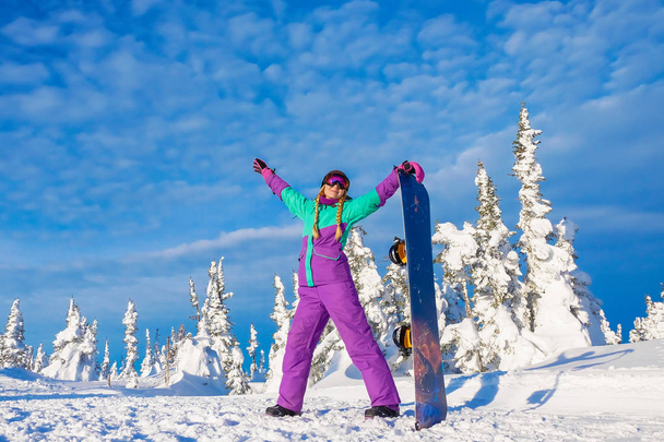 Successful young girl snowboarding in the mountains Sheregesh. Snowboarder resting.  Stylish caucasian snowboarder. Snowboarder smiling at the camera on top of the mountain on a background of sky.  - Photo, Image
