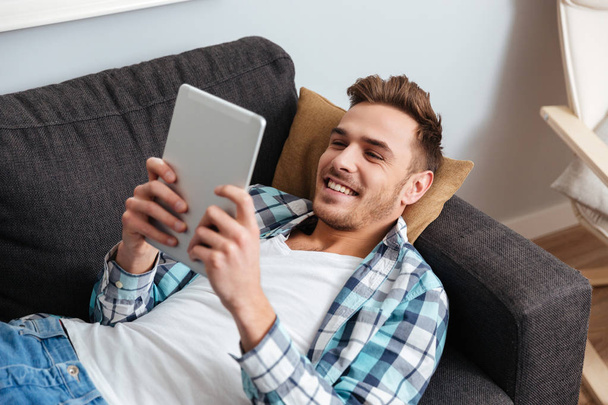 Cheerful bristle man lies on sofa and using tablet computer - Foto, Imagem