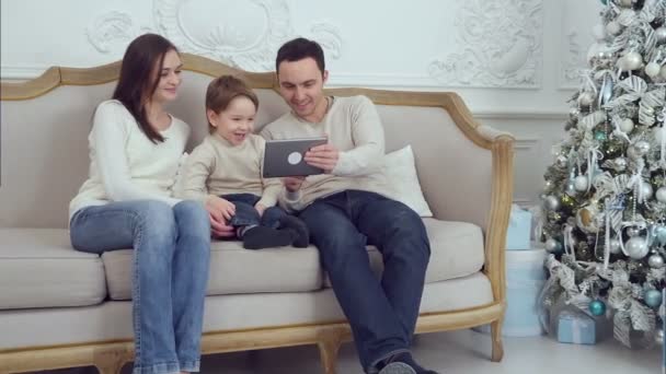 Dad, mom and their little son having fun by playing together with a tablet sitting on a couch - Кадри, відео