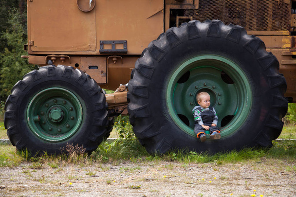 Małe słodkie dziecko siedzi w ogromne koła ciągnika. Maszyna wyciągowa drewna. - Zdjęcie, obraz