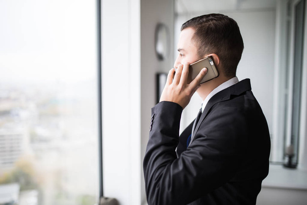 Handsome young businessman standing next to the large windows of his top floor office, looking at the view of the city while talking on his mobile phone - 写真・画像