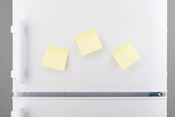 Three blank yellow sticky paper notes on white refrigerator - Foto, afbeelding