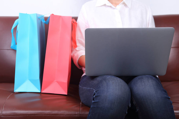 женщина сидит на диване с Shoping сумка рядом и с помощью ноутбука - Фото, изображение