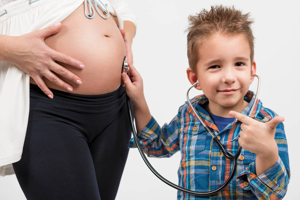 Lapsen ja raskaana olevan naisen muotokuva. Poika käyttää stetoskooppia. Hän kuuntelee, mitä äitini vatsassa tapahtuu. Valokuvan lähikuva
 - Valokuva, kuva