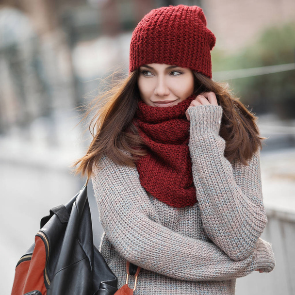 Πορτρέτο του όμορφη κοπέλα με ένα ζεστό πουλόβερ, ένα κόκκινο σκουφάκι και ένα μαντήλι και ένα σακίδιο στον ώμο του - Φωτογραφία, εικόνα