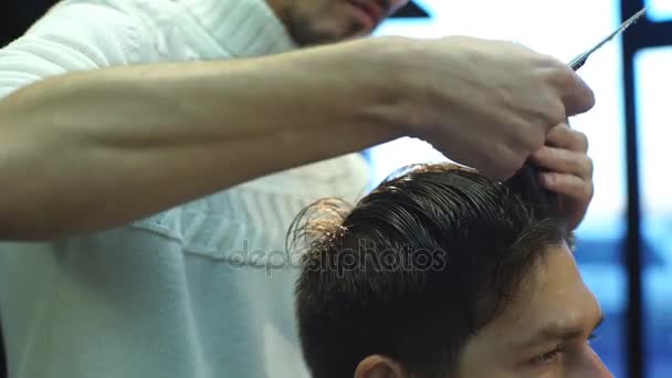 Young bearded man getting haircut by hairdresser while sitting in chair at barbershop - Felvétel, videó