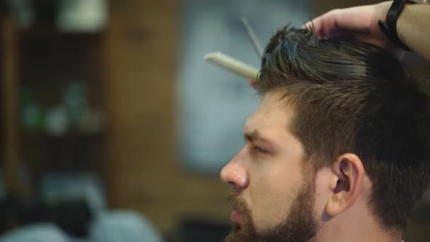 Young bearded man getting haircut by hairdresser while sitting in chair at barbershop - Záběry, video