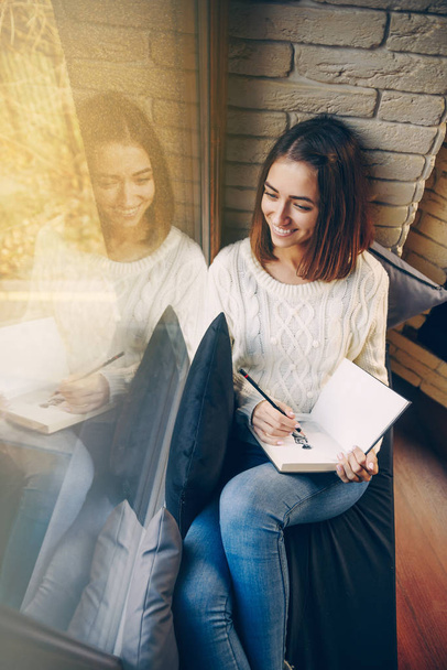 Young beautiful smiling girl in a white sweater and jeans sitting on the window sill and drawing a picture in sketchbook - Foto, Bild