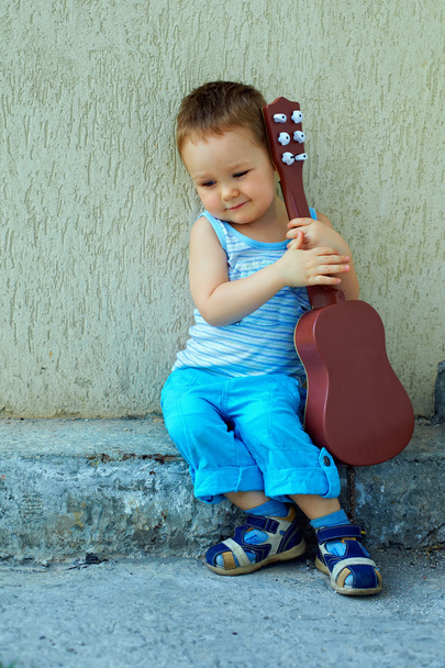 Cute baby boy with guitar sitting against the concrete wall - Foto, afbeelding