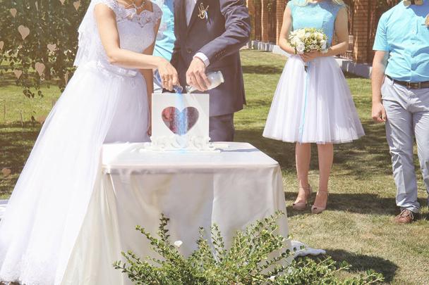 Groom and bride pouring colourful sand into box with glass heart outdoor - Zdjęcie, obraz