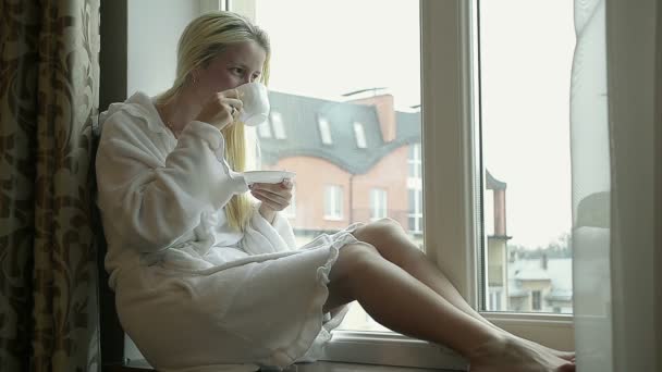 Chica en Albornoz Beber café en casa cerca de la ventana y mirando hacia fuera
  - Imágenes, Vídeo