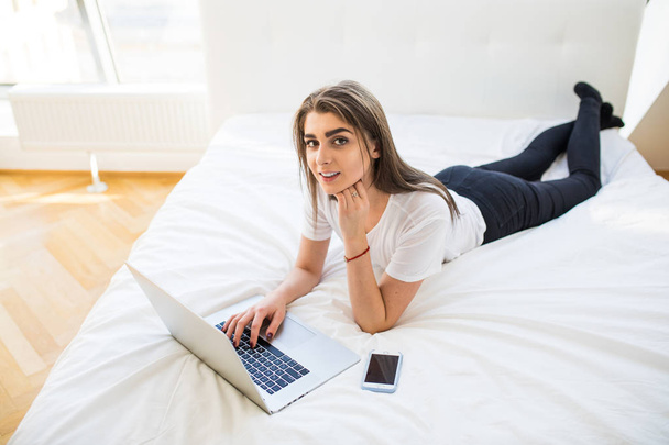 Portrait of beautiful happy young woman lying in bed and using laptop at home - Photo, Image