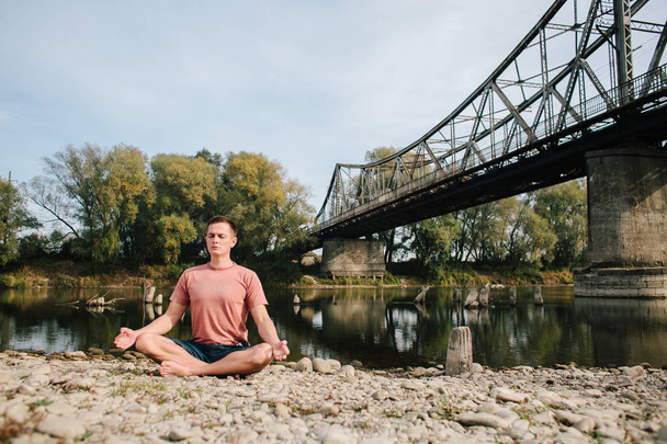 man practices yoga on the river bank near the old bridge. lotos - Photo, Image