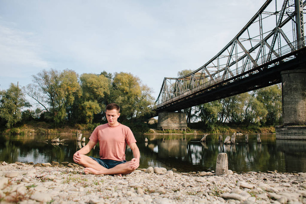 man practices yoga on the river bank near the old bridge - Photo, Image
