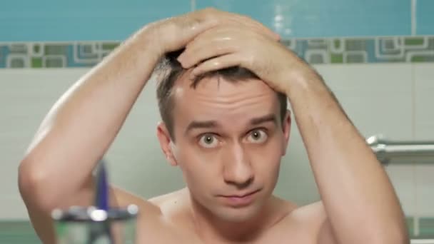 Young attractive man straightens hairstyle after a shower in the bathroom of the hotel room. He hands combs hair in the desired direction. He is looking at the camera in the mirror. - Materiaali, video