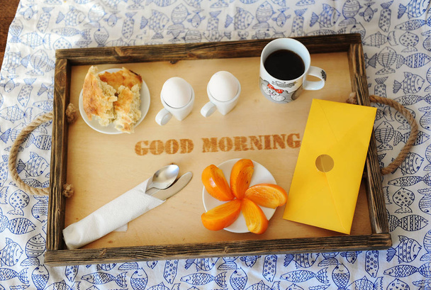 Ontbijt met eieren en koffie op een houten dienblad met opschrift "good morning". Tafelkleed met vis. Gele envelop - Foto, afbeelding