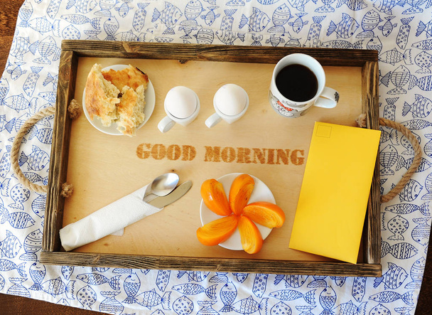 Desayuno con huevos y café en una bandeja de madera con inscripción "buenos días". Mantel con pescado. Sobre amarillo
 - Foto, Imagen