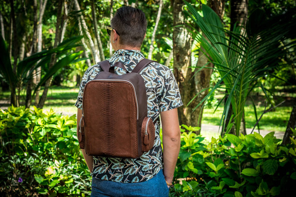 Tourist with stylish brown snakeskin python backpack in the asian park. Bali, Indonesia. Handsome caucasian man in sunglasses. Rare view. - Photo, Image