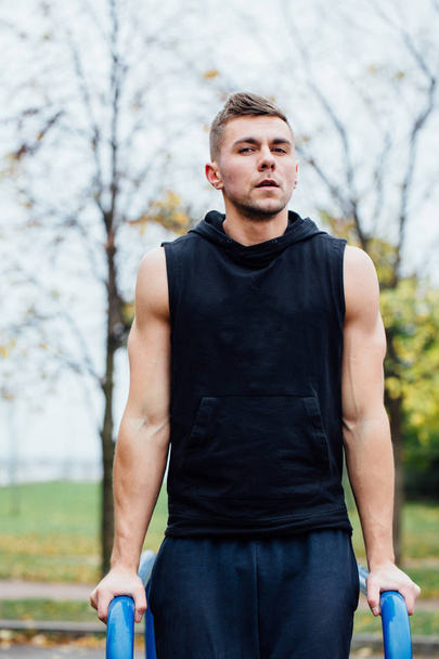 портрет сфокусированного мускулистого молодого человека в черной тренировочной одежде, делающего погружения на параллельных брусьях
. - Фото, изображение