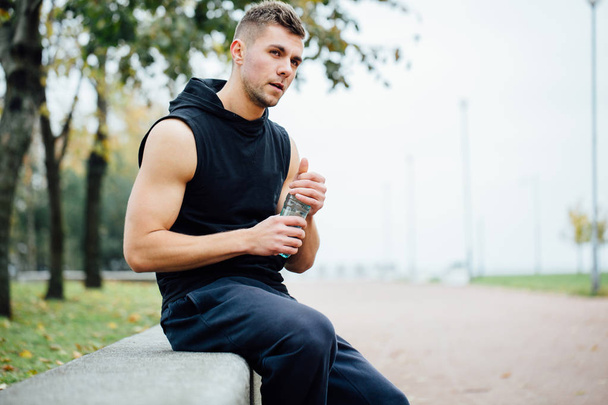 Athlete resting on bench in park after running with bottle of water. Rest  a hard workout. - Zdjęcie, obraz