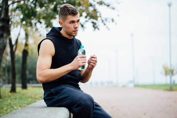 Athlete resting on bench in park after running with bottle of water. Rest  a hard workout. - Zdjęcie, obraz