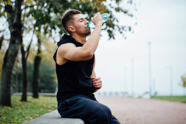 Athlete resting on bench in park after running with bottle of water. Rest  a hard workout. - Foto, immagini