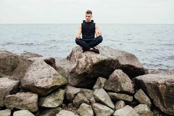 Strong fitness yoga man in lotus pose on the rock beach near the ocean. Harmonic concept. - Foto, afbeelding