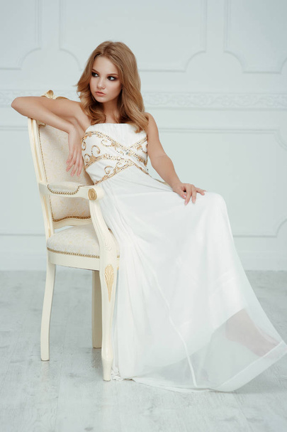 Smartly dressed charming young beautiful girl (teenager) with frail figure and blond wavy hair wearing white evening dress embroidered with sequins is posing in the light interior studio room  - Photo, Image