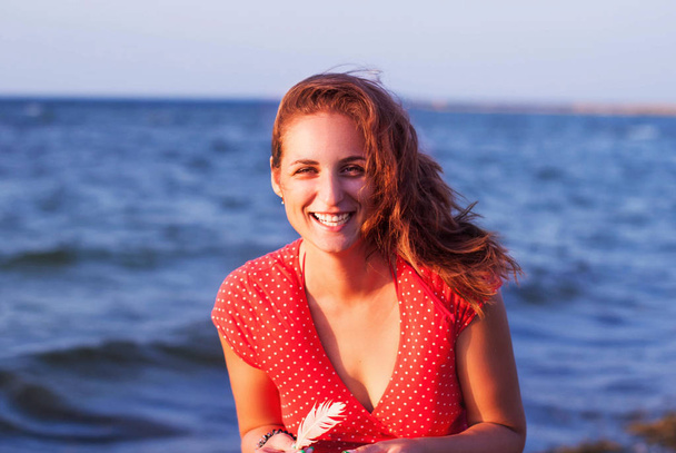Jong meisje in een rode jurk glimlachend op de achtergrond van de zee - Foto, afbeelding