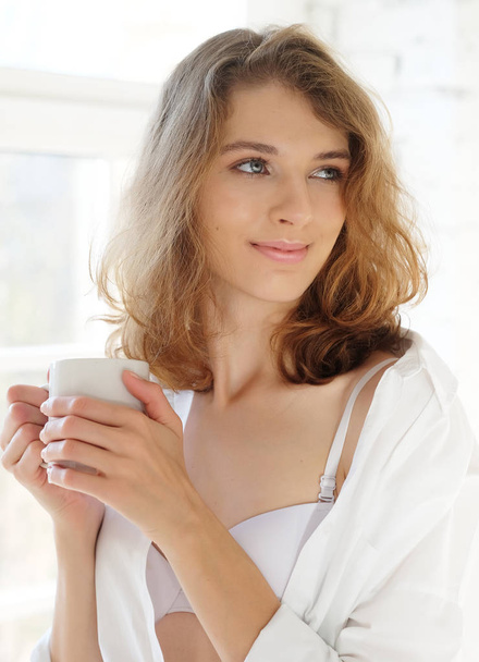 Beautiful woman wearing white lingerie drinking coffee in the mo - Фото, изображение