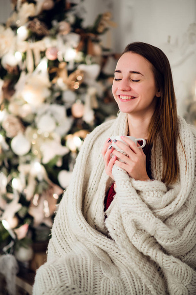 Счастливая молодая женщина с чашкой горячего напитка возле рождественской елки - Фото, изображение